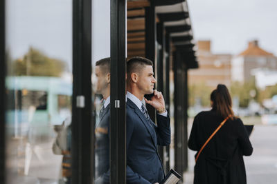 Businessman talking on smart phone while leaning on glass door at street