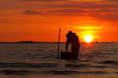 Silhouette man standing on shore against orange sunset sky