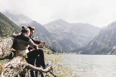 Austria, tyrol, alps, couple relaxing on tree trunk at mountain lake using cell phone