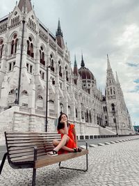 Full length of woman sitting outside hungarian parliament building on bench in city