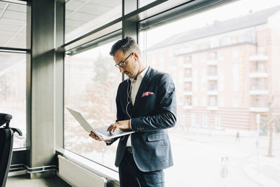 Mature businessman using laptop while standing against window in office