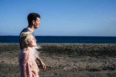 Side view of couple standing at beach against blue sky