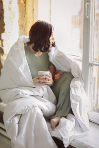 Young woman holding coffee cup sitting by window