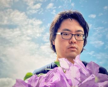 Portrait of young man behind purple clematis flowers against cloudy blue sky.