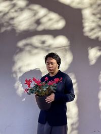 Portrait of young asian man holding red flowering cyclamen plant against shadow patterned wall.