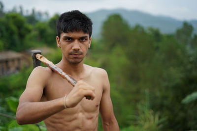 Portrait of shirtless young man holding axe standing outdoors