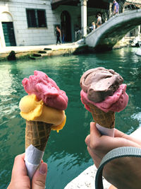 Cropped image of hand holding ice cream cone against canal