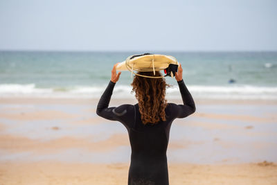Rear view of man holding surfboard while standing at beach against sky