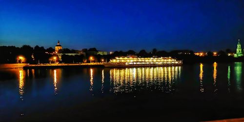 Illuminated buildings by lake against blue sky at night