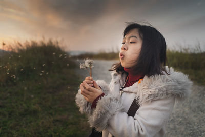 Young woman blowing dandelion on land against sky during sunset