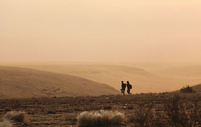 Man and woman walking on field against sky during sunset