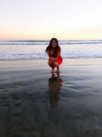 Full length of happy woman crouching at beach against sky during sunset