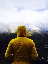 Rear view of person in yellow hooded jacket standing against snowcapped mountains