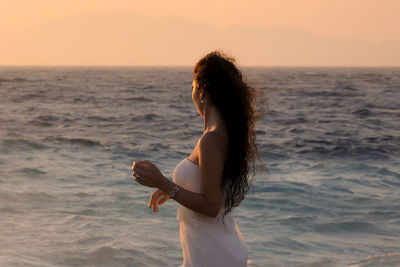 Side view of woman standing by sea against sky during sunset