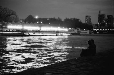 Woman sitting on illuminated city by sea against sky at night