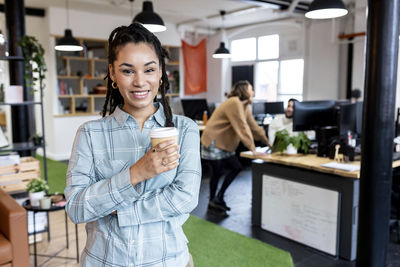 Happy businesswoman standing with disposable cup in office