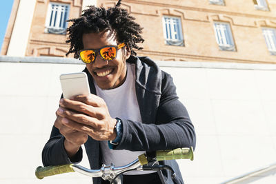 Low angle view of smiling young man wearing sunglasses using mobile phone against building