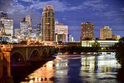 Bridge over river by illuminated buildings against sky at night