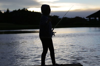 Boy standing on riverbank against sky during sunset