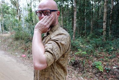 Close-up of man with hand covering mouth standing on street against trees