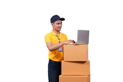 Smiling delivery man using laptop by cardboard boxes against white background