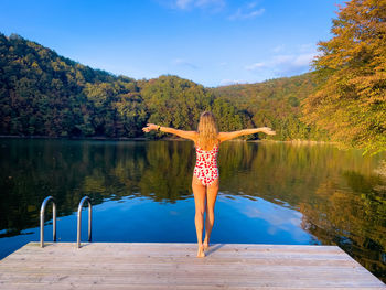 Rear view of young woman in swimming suit standing on a wooden pontoon near a lake 