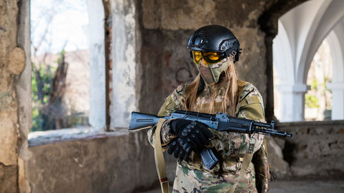 A woman in an army uniform aims to shoot a firearm in an abandoned building. 