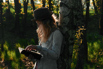 Side view of woman reading book by trees in forest