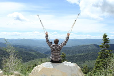 Rear view of man holding crutches while sitting on rock against mountains