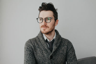 A young stylish man with a beard and glasses in a shirt and a gray cardigan against a gray wall. 