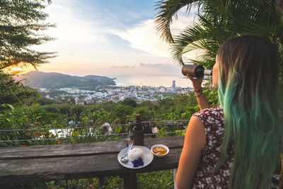 Rear view of young woman drinking by table against sky during sunset