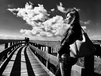 Side view of young woman looking away while standing on footpath against sky