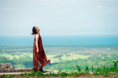 Side view of young woman wearing sunglasses standing on observation point against cloudy sky