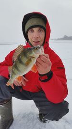Portrait of mature man holding fish while crouching on snow covered land