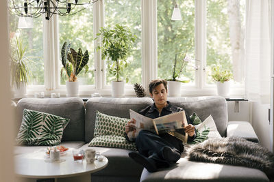 Young man reading newspaper while sitting on sofa in living room at home