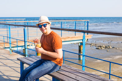 Man sitting on railing by sea against sky