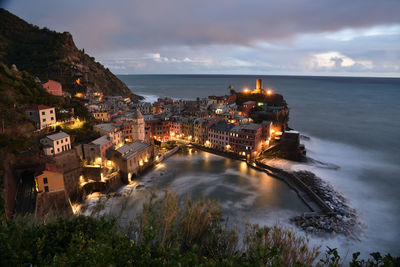 Illuminated buildings by sea against sky at dusk