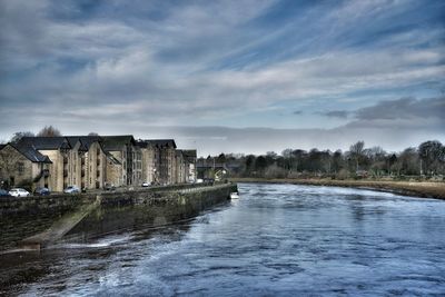 Buildings by river against cloudy sky