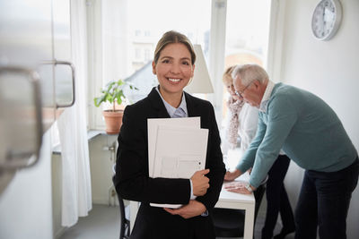 Portrait of smiling real estate agent holding documents with senior couple in background