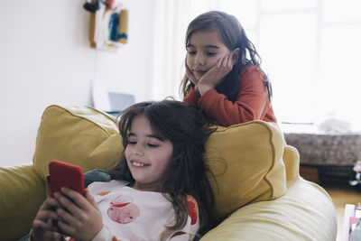 Girl with head in hands looking at sister using phone while sitting on armchair in living room