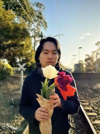 Young asian man holding a bunch of tulips at railroad crossing against trees and sunset sky