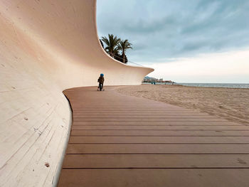 Rear view of child riding his bike on a boardwalk by the beach against sky