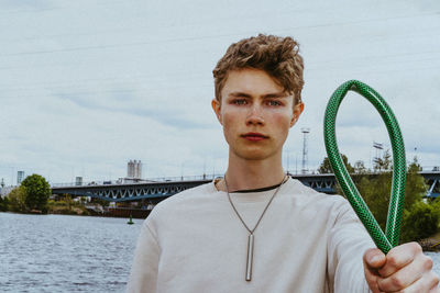 Portrait of confident man holding garden hose near river against sky