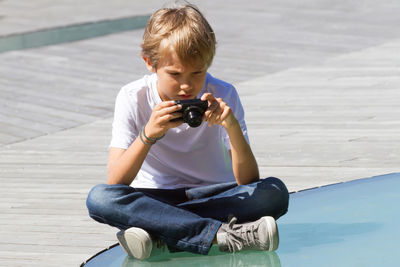 Boy photographing while sitting on footpath in city