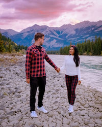 Couple holding hands while standing at riverbank against mountains