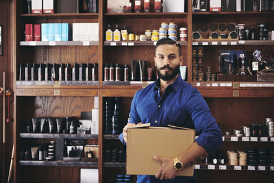 Portrait of confident salesman carrying box while standing against rack in deli