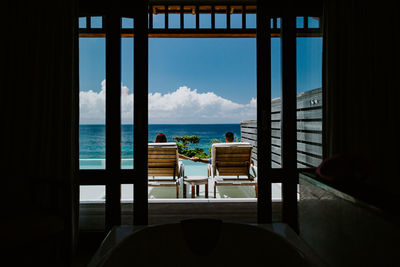 Rear view of couple holding hands while sitting on lounge chair at poolside by sea