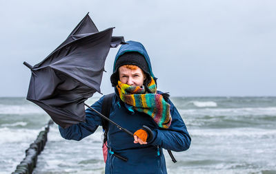 Portrait of woman holding damaged umbrella while standing at shore of beach against sky