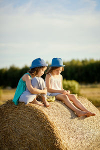 Friends wearing hats sitting on hay bale against sky