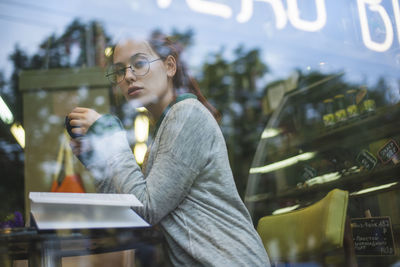 Through glass side view of relaxed female in casual outfit sitting at table with opened book in cozy coffee shop and enjoying aromatic beverage while looking at camera